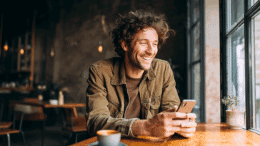 Jeune personne souriante utilisant un iPhone reconditionné dans un café, avec une tasse de café et une ambiance chaleureuse.